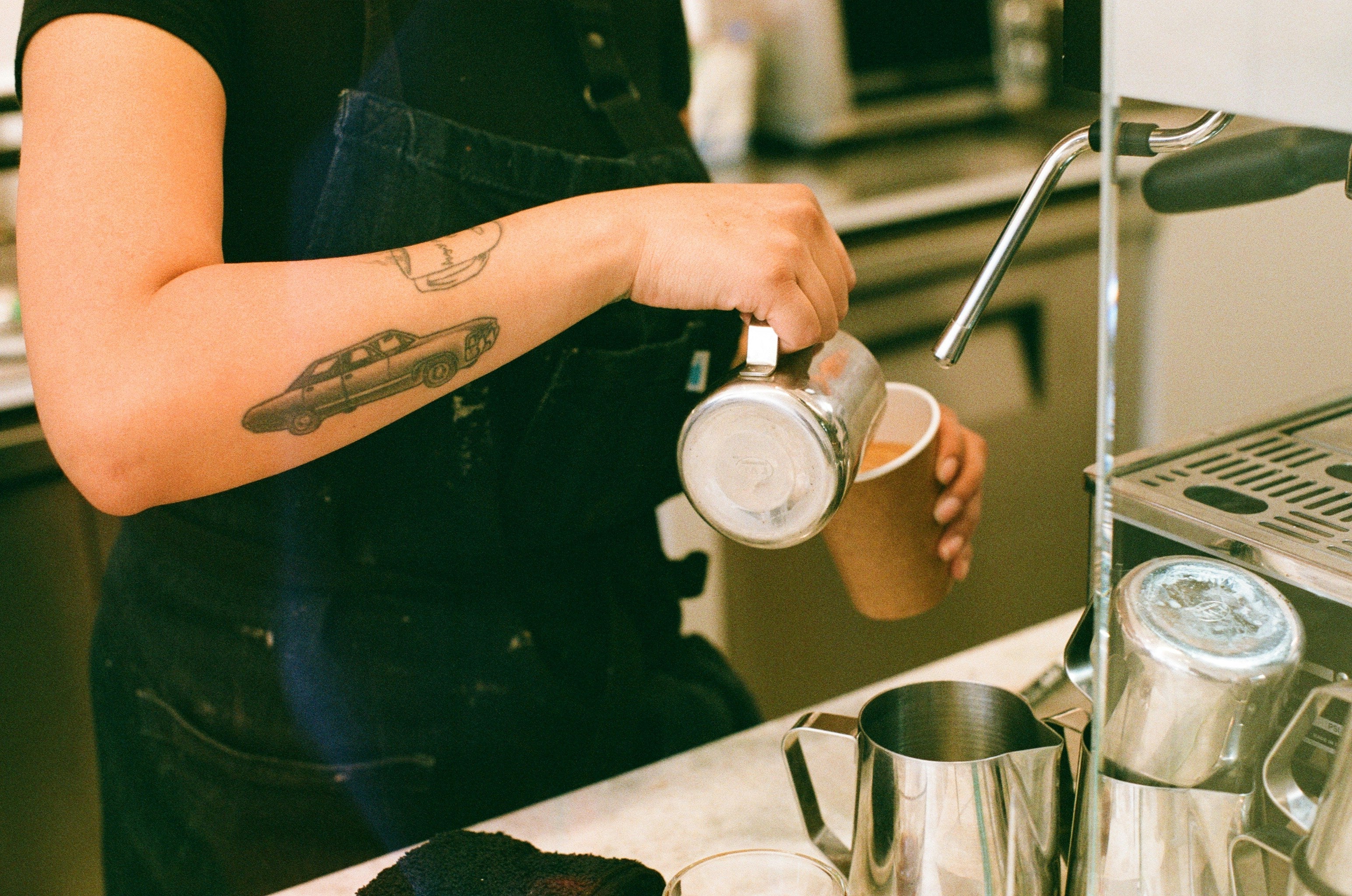 scene of barista pouring coffee, tattoos on arm of a car and hot cup of coffee